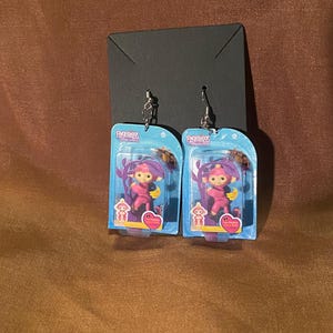Puede incluir: Un par de pendientes con personajes Fingerlings rosas en un embalaje de plástico transparente. El embalaje tiene el logotipo de Fingerlings y las palabras "Love, Friendship, and Fun!" impresas en él.