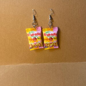 May include: A pair of earrings featuring miniature Fruit-tella candy bags. The bags are yellow with pink trim and feature a picture of assorted fruit slices. The earrings are silver and have a hook closure.