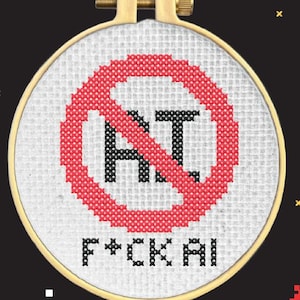 Puede incluir: Un bastidor de bordado circular de madera que contiene tela Aida blanca con un diseño de punto de cruz. El diseño presenta un símbolo de prohibición circular rojo sobre las letras negras AI, con el texto negro F*CK AI bordado debajo.