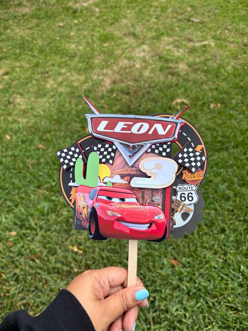 May include: A red and black Cars-themed cake topper with the name "LEON" and the number "3". The design includes Lightning McQueen, Route 66 imagery, and checkered flags, all attached to a wooden stick.