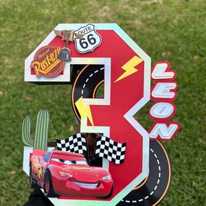May include: A red and white number three birthday decoration featuring Lightning McQueen from the Cars movie. The decoration includes a Route 66 sign, a trophy, and the name "LEON".