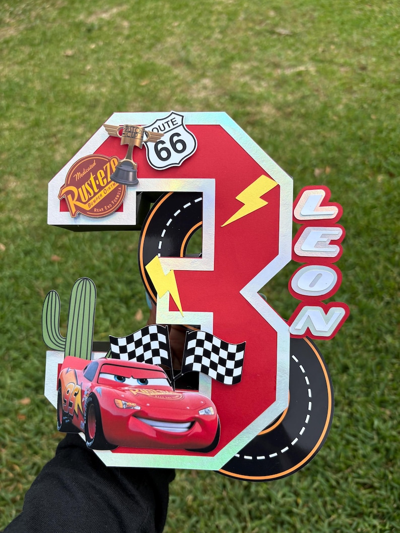 May include: A red and white number three birthday decoration featuring Lightning McQueen from the Cars movie. The decoration includes a Route 66 sign, a trophy, and the name "LEON".