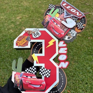 May include: A red and white number 3 cake topper featuring Lightning McQueen from the Cars movie. The topper includes the name "LEON", Route 66 signs, checkered flags, and a cactus. The background is green.