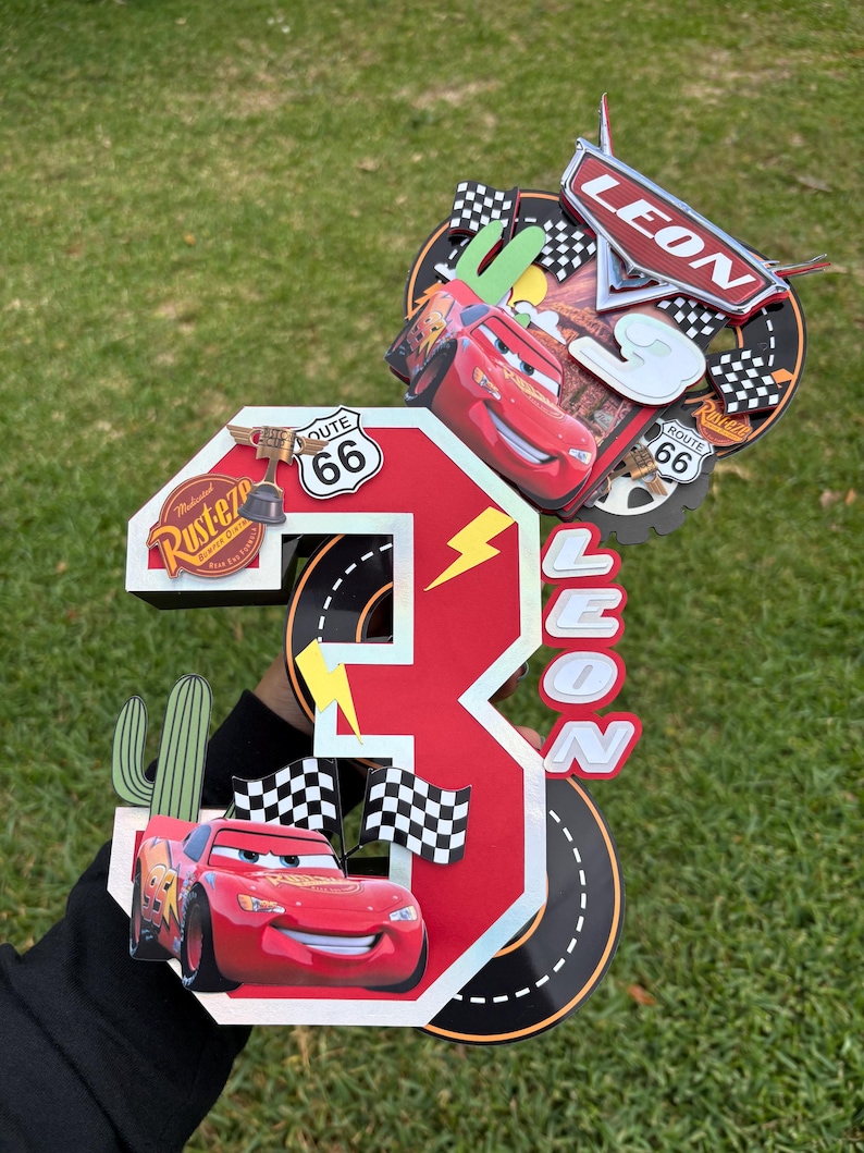 May include: A red and white number 3 cake topper featuring Lightning McQueen from the Cars movie. The topper includes the name "LEON", Route 66 signs, checkered flags, and a cactus. The background is green.
