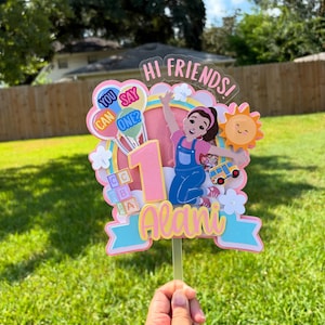 May include: A colorful cake topper with the text "Hi Friends!" and "You Can Say One?" The topper features a cartoon girl, a number 1, a sun, and the name "Alani". The design includes balloons, flowers, and a toy bus.