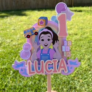 May include: A decorative cake topper featuring a cartoon character with brown hair and a pink headband, wearing a pink shirt and blue overalls. It includes a yellow school bus, a rainbow, a pink number one, alphabet blocks, and the name LUCIA.
