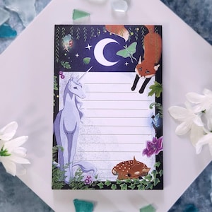 May include: A notepad with a white background and lined paper. The notepad has a black border and features illustrations of a unicorn, a fox, a deer, and a crescent moon. The illustrations are surrounded by green leaves and flowers.