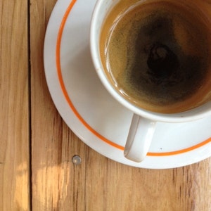 May include: A white ceramic coffee cup with an orange rim, filled with dark coffee, sitting on a white saucer with an orange rim. The cup and saucer are on a wooden surface.