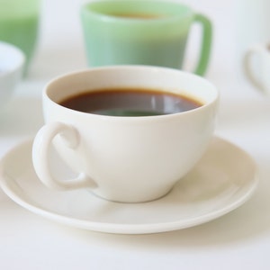 May include: A white coffee cup with a saucer, filled with dark coffee. The cup is on a white surface with other cups and saucers out of focus in the background.