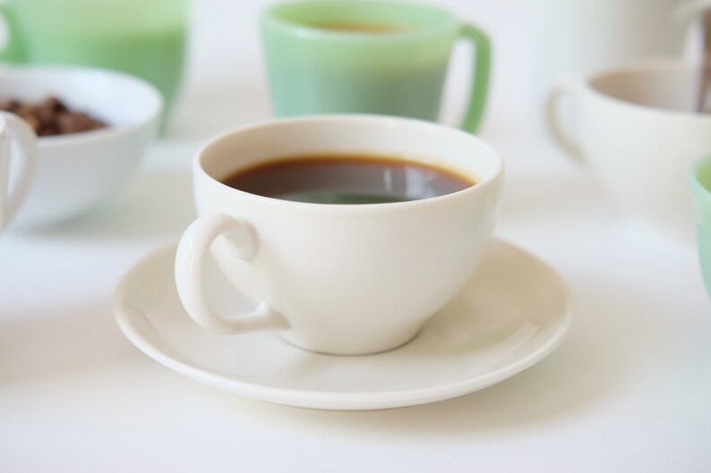 May include: A white coffee cup with a saucer, filled with dark coffee. The cup is on a white surface with other cups and saucers out of focus in the background.