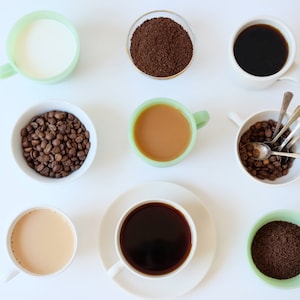 May include: A flat lay of coffee making supplies, including a cup of milk, a cup of coffee, a cup of creamer, a bowl of coffee beans, a bowl of ground coffee, and a cup of coffee grounds.