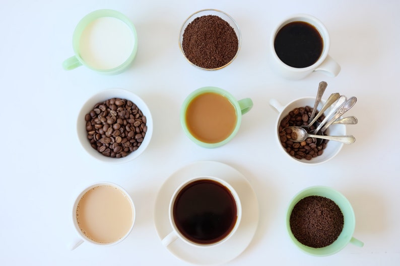 May include: A flat lay of coffee making supplies, including a cup of milk, a cup of coffee, a cup of creamer, a bowl of coffee beans, a bowl of ground coffee, and a cup of coffee grounds.