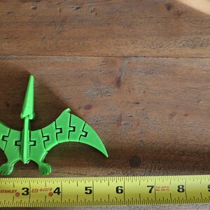 May include: A green plastic pterodactyl toy, approximately 4 inches long, is positioned on a wooden surface. A yellow measuring tape is visible at the bottom of the image.
