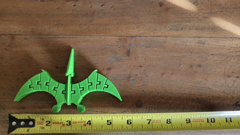 May include: A green plastic pterodactyl toy, approximately 4 inches long, is positioned on a wooden surface. A yellow measuring tape is visible at the bottom of the image.