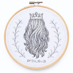 Può includere: Un telaio da ricamo circolare in legno contenente tessuto Aida bianco con un disegno a punto croce in filo nero. Il motivo raffigura una corona sopra lunghi capelli ondulati, fiancheggiata da due rami frondosi, con il testo PROV. 16:31.