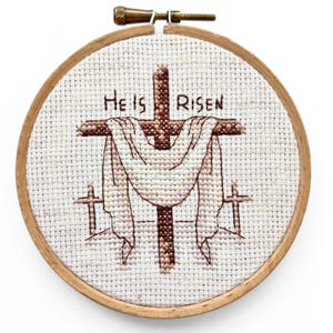 May include: A cross-stitch embroidery in a wooden hoop. The design features a brown cross with a draped white cloth and the words "He Is Risen" stitched above. Two smaller crosses are on either side.