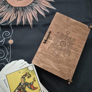 May include: A wooden tarot card box with a sun and moon design, a metal clasp, and a deck of tarot cards. The cards feature colourful illustrations. The background is a black cloth with a sun and moon design.