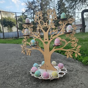 May include: A wooden Easter tree with laser-cut floral designs, holding painted Easter eggs on small circular shelves. The tree has a heart-shaped cutout and sits on a decorative base with more eggs. The eggs are pastel colors.
