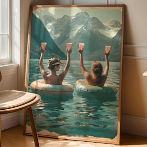 Könnte beinhalten: Ein gerahmter Kunstdruck zeigt zwei Personen in Schwimmreifen in einem See, die rosa Getränke hochhalten. Der Hintergrund zeigt eine Bergkette und eine ruhige See-Szene. Das Kunstwerk wird neben einem Fenster und einem Holzstuhl ausgestellt.