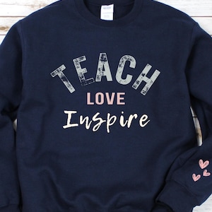 Op de afbeelding: Donkerblauwe trui met de tekst "Teach Love Inspire" in wit en roze. De trui heeft roze hartjes op de manchetten.