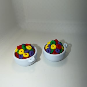 May include: Two small white ceramic bowls, each filled with colorful, ring-shaped cereal. The cereal pieces are red, yellow, green, and purple. The bowls have small handles on either side.