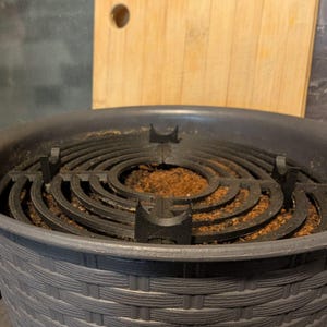 Puede incluir: Una maceta gris oscuro, con textura tejida, con un inserto circular negro. El inserto tiene anillos concéntricos y soportes, llenos de tierra marrón. Una tabla de cortar de madera está en el fondo.