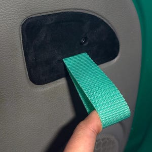 May include: Close-up of a green fabric pull strap attached to a gray car door panel. The strap is held by a finger. The panel has a black, oval-shaped insert.