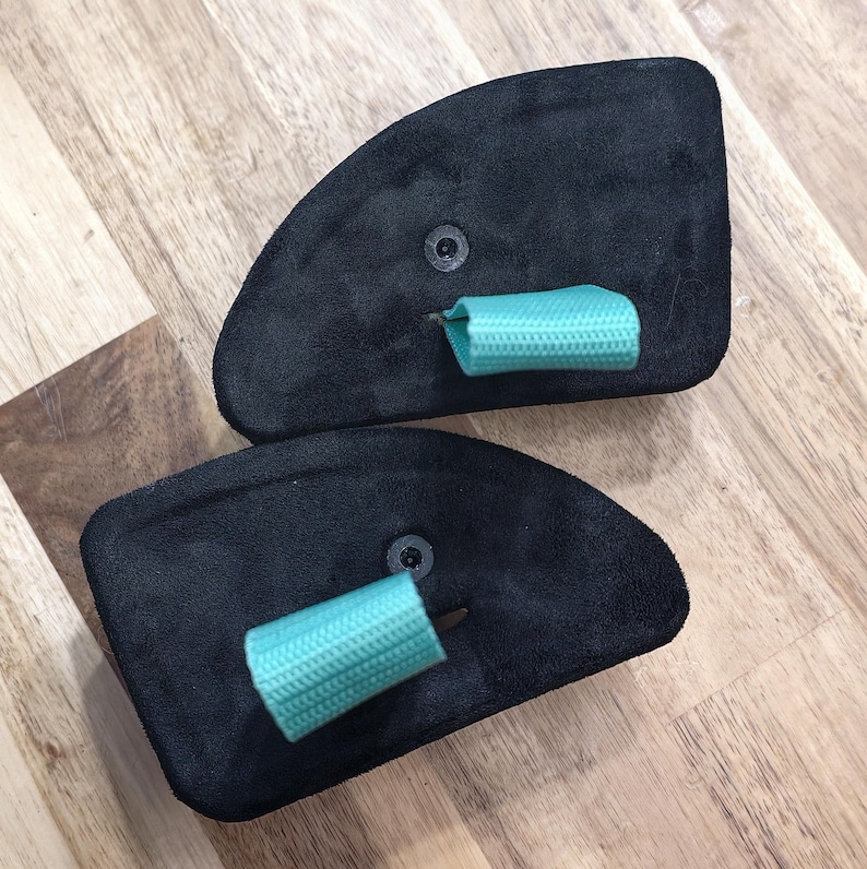 May include: Two black suede-like pads with a rounded shape, each featuring a small, light blue fabric loop. The pads are secured with a small, dark fastener. The items are displayed on a light brown wooden surface.