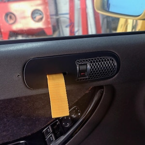 Könnte beinhalten: Ein schwarzer Ersatz-Türgriff für ein Auto mit einem gelben Nylon-Zugband und einem strukturierten Lautsprechergitter im Wabenmuster. Die Baugruppe ist auf einer dunkelgrauen Innentürverkleidung über den Fenster- und Spiegelsteuerungen montiert.