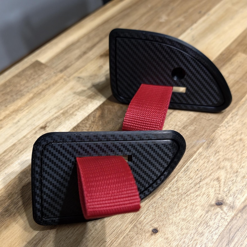 May include: Two black carbon fiber car door pulls with red nylon straps. The pulls have a textured surface and a curved design. The straps are visible, extending from the pulls.
