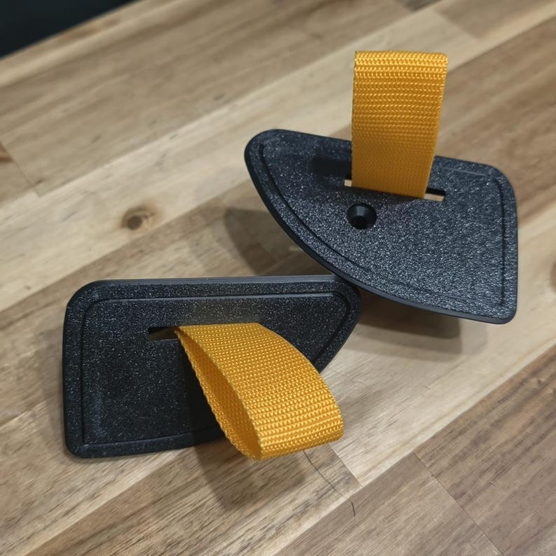 May include: Two black, textured, plastic door pulls with bright yellow fabric loops. The pulls are angled and have a small hole and slot for the fabric. The items are on a wooden surface.