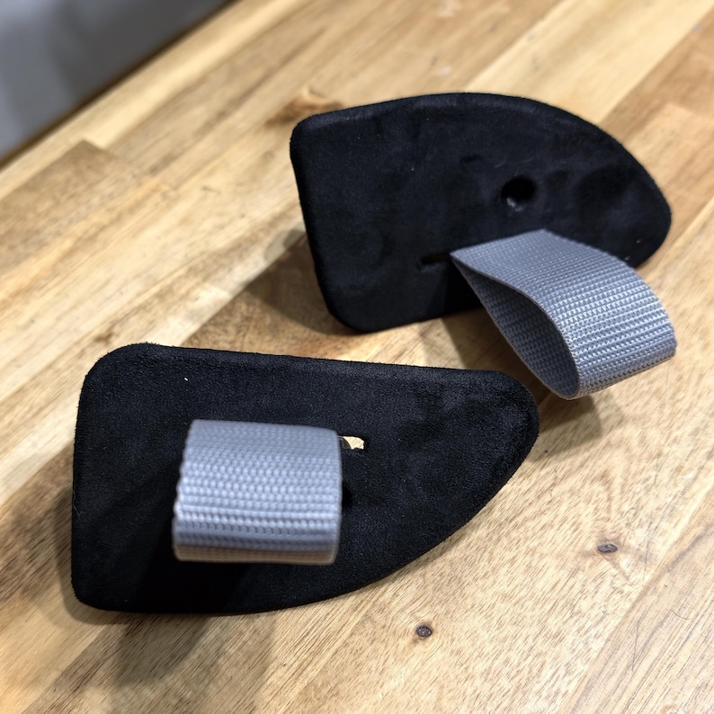 May include: Two black climbing holds with gray webbing loops are displayed on a wooden surface. The holds have a curved shape and a textured surface. The gray loops are attached to the holds.