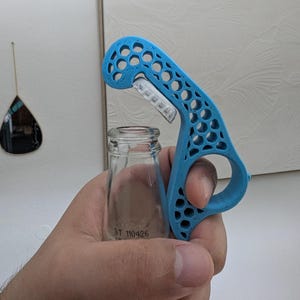 May include: A blue bottle opener with a honeycomb design is held over a small glass bottle. The opener has a curved handle with a finger hole and a metal blade. The bottle has the text "DT 110426" printed on it.