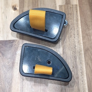 May include: Two black, semi-circular objects with a yellow strap. The objects have a dark gray border and are set against a light brown wooden surface. One object has a metal hook.