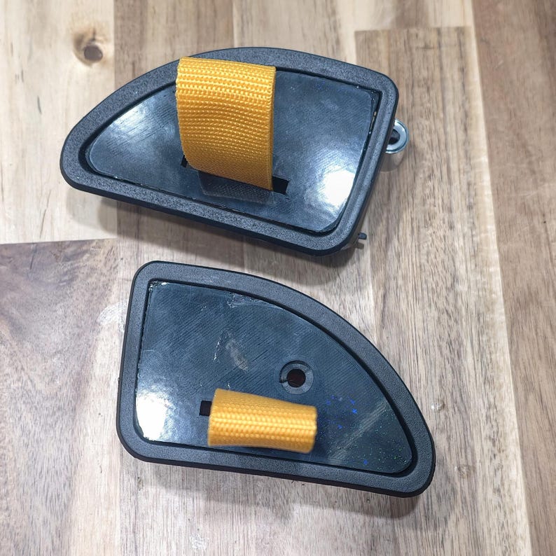 May include: Two black, semi-circular objects with a yellow strap. The objects have a dark gray border and are set against a light brown wooden surface. One object has a metal hook.