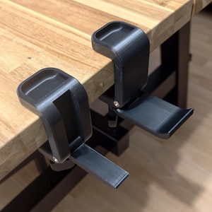 May include: Two black clamps are attached to a wooden surface. Each clamp has a curved top section with a small rectangular indentation and a hinged, flat base. The clamps appear to be made of a matte black plastic material.