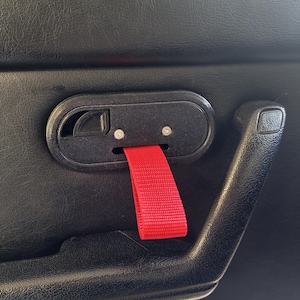 Peut inclure: Un panneau de porte de voiture intérieur noir doté d'un mécanisme d'ouverture ovale noir avec deux petites vis argentées et une sangle de traction en tissu rouge vif orientée vers le bas, à côté d'une poignée en plastique noir texturé.
