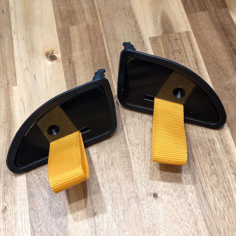 May include: Two black car interior handles with bright yellow fabric loops. The handles have a curved design and are set against a wooden surface. The yellow loops are made of a woven material.