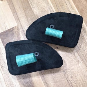 May include: Two black suede-like pads with a curved shape, each featuring a small, cylindrical, light teal-colored grip. The pads are positioned on a light brown wooden surface, creating a contrasting color scheme.