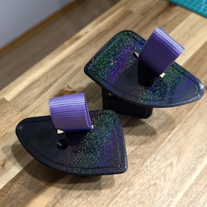 May include: Two black, angled objects with a holographic, speckled surface and a purple fabric loop. The objects are on a wooden surface. The fabric loops are a light purple color.