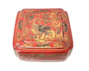 May include: A red, square-shaped decorative box with a lid. The box is adorned with gold and black ornate designs, including a dragon and cloud motifs. The box has a glossy finish and is set against a white background.