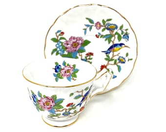 May include: A white teacup and saucer with a floral and bird design. The cup and saucer have a gold trim. The floral design features pink, blue, yellow, and green flowers. The bird is blue with a yellow beak and a red breast.