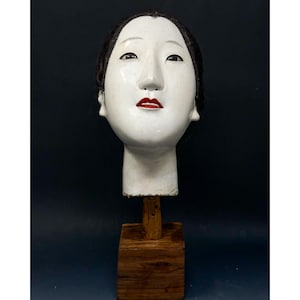 May include: A white, stylized face with black hair and red lips, mounted on a wooden stand. The face has a serene expression and is likely a decorative art piece or sculpture.