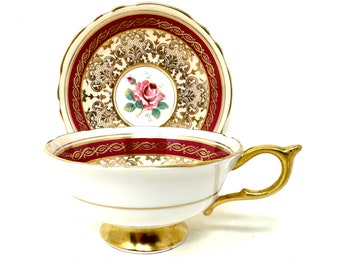May include: A white teacup and saucer with a gold rim and a red and gold floral design. The saucer has a scalloped edge.