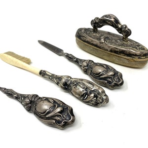 May include: A vintage silver-plated vanity set with a brush, two nail tools, and a compact. The tools have ornate floral designs.