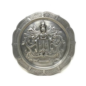 May include: A silver pewter plate with a raised design of a coat of arms featuring a crown, a shield with the letters "L.I.G." and a banner with the word "LIEGE".