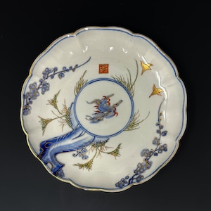 May include: A white ceramic plate with a scalloped edge and gold trim. The plate features a central circular design with a blue bird and red accents, surrounded by green and gold foliage. Blue floral and gold bird accents are also present.
