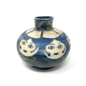 May include: A small, blue and white ceramic vase with a rounded body and a wide mouth. The vase is decorated with white stylized faces and geometric patterns.