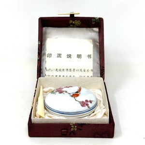 May include: A small, round, porcelain inkwell with a lid. The lid is decorated with a red bird perched on a branch of pink blossoms. The inkwell is in a red box with a floral pattern. The box contains a small, ivory-colored stick. The box has a label that reads "印泥说明书" in Chinese characters.