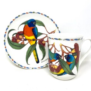 May include: A white ceramic teacup and saucer set with a colorful bird design. The saucer features a single bird with blue, orange, and yellow feathers perched on a branch with green leaves and red flowers. The teacup features two birds with blue, green, and yellow feathers perched on branches with green leaves and red flowers. The text "Scents of Chester" is printed on the saucer.